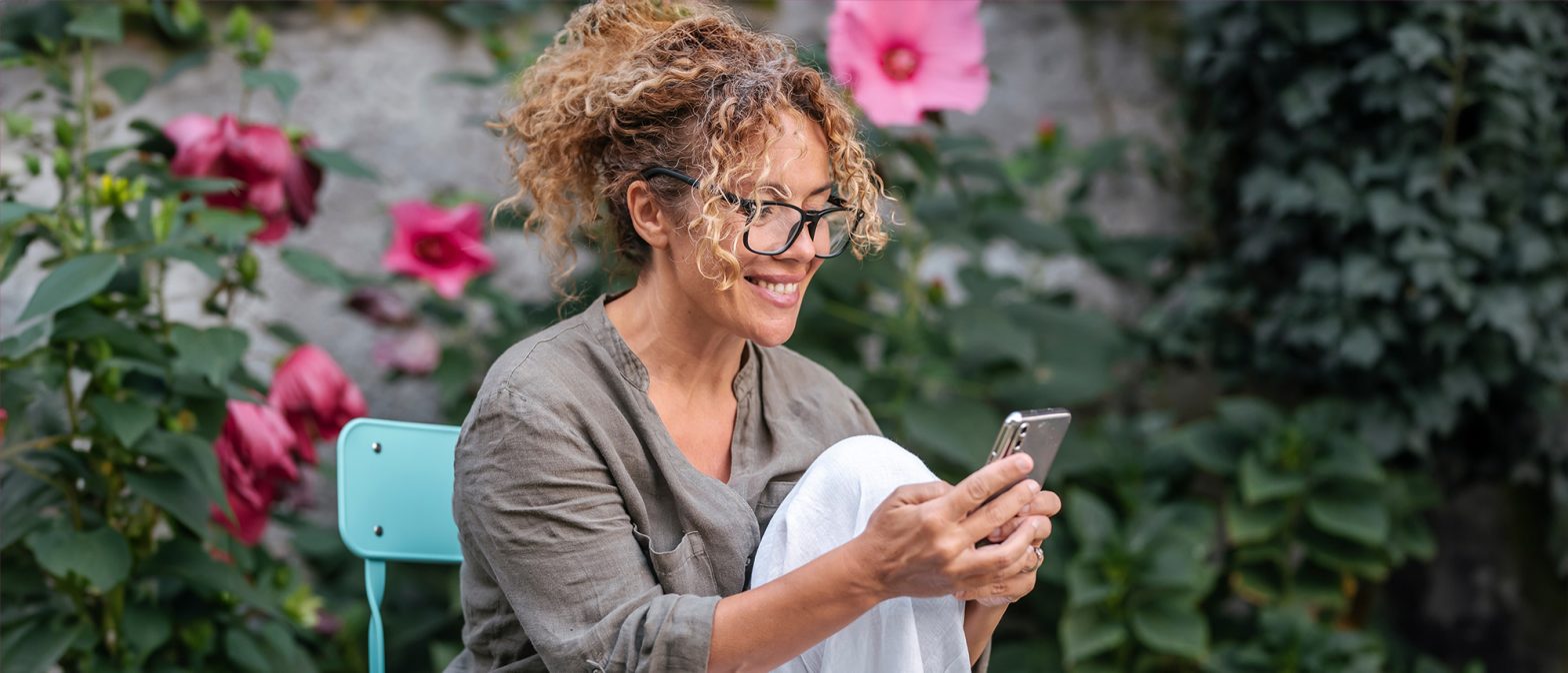 En kvinne med krøllete hår og briller sitter smilende på en blå stol utendørs mens hun ser på sin håndholdte smarttelefon. Hun er kledd i en avslappet grå skjorte og hvite bukser. Bak henne er det uskarpe grønne planter og rosa blomster som gir en rolig og frodig bakgrunn.