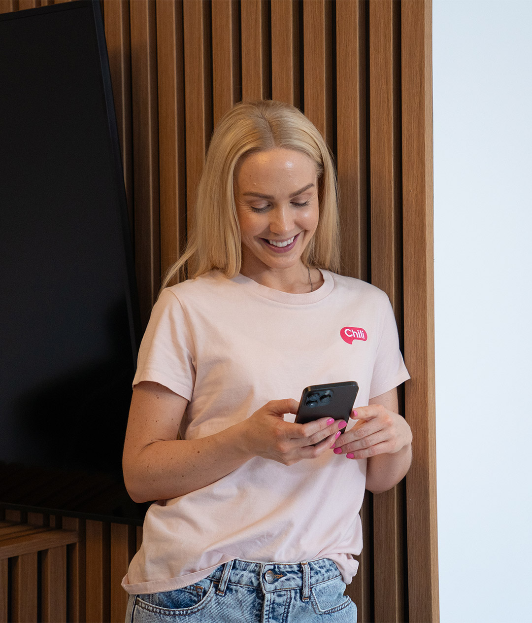 Nærbilde av en kvinne med blondt hår som står ved en vegg med trepanel og ser ned på mobilen. Hun smiler og har på seg en rosa T-skjorte med teksten “Chill” og lyse dongeribukser