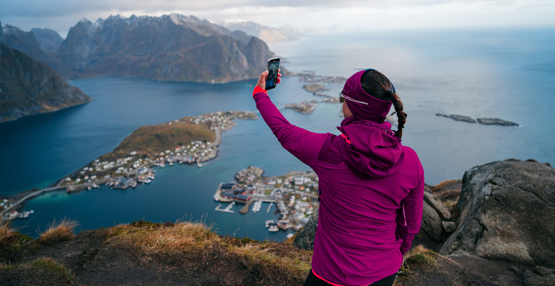 Kvinne står på toppen av et fjell, med en mobil og ser utover en norsk by