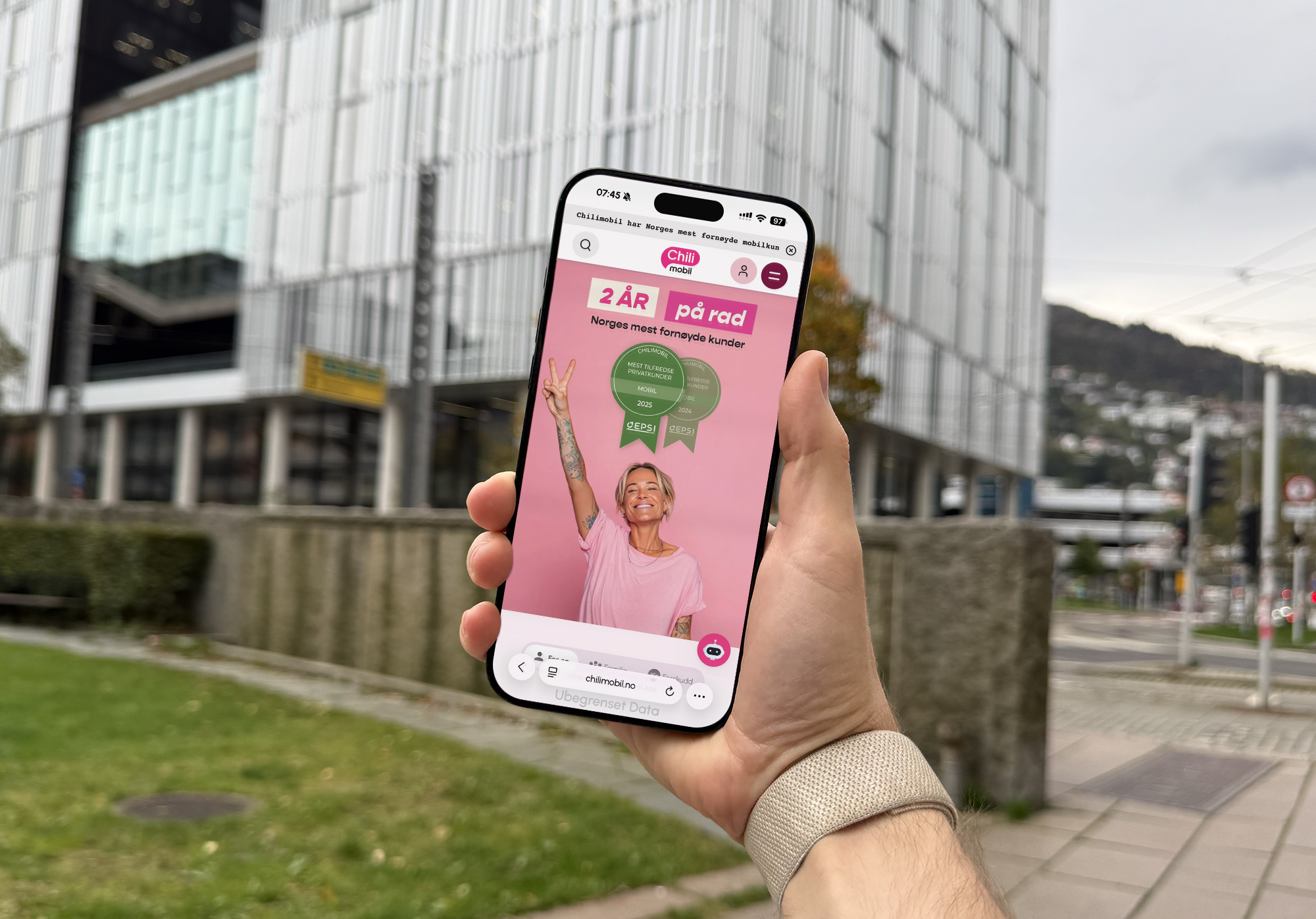 En hånd holder en smarttelefon ute i bymiljø, med en nettside åpen som viser forsiden til Chilimobil. På skjermen vises en smilende person i rosa t-skjorte som gjør fredstegn foran rosa bakgrunn, sammen med teksten «2 år på rad – Norges mest fornøyde kunder» og EPSI-utmerkelser for 2024 og 2025.